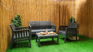 Un patio con un muro de madera, una banca, dos sillas, una mesita con un vaso y luces colgantes.