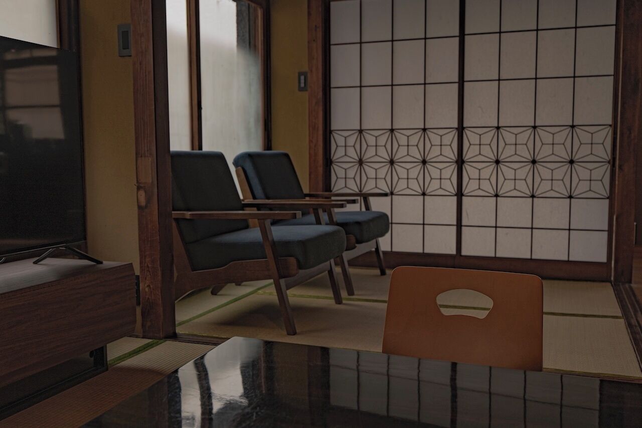 A traditional Japanese room with tatami flooring, a low wooden table, two dark blue chairs, a television, and a shoji screen.