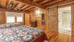 A log cabin bedroom with two beds, a ceiling fan, a large sliding door, and a window with curtains. - Dragonfly Meadows Log Cabin (Stoutland)