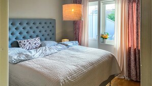 A bedroom with a tufted headboard, a bed with patterned bedding, a bedside lamp, and a window with curtains. - Amazing apartment in Färjestaden (Färjestaden)