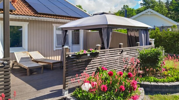 A wooden deck with two lounge chairs, a covered patio area, and a flower bed with pink flowers. - Amazing apartment in Färjestaden (Färjestaden)
