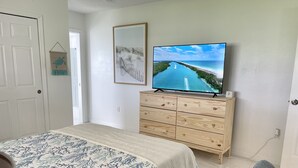 Ein Schlafzimmer mit einem Bett, einer hölzernen Kommode, einem Fernseher, der eine Strandansicht zeigt, und einem gerahmten Bild an der Wand.