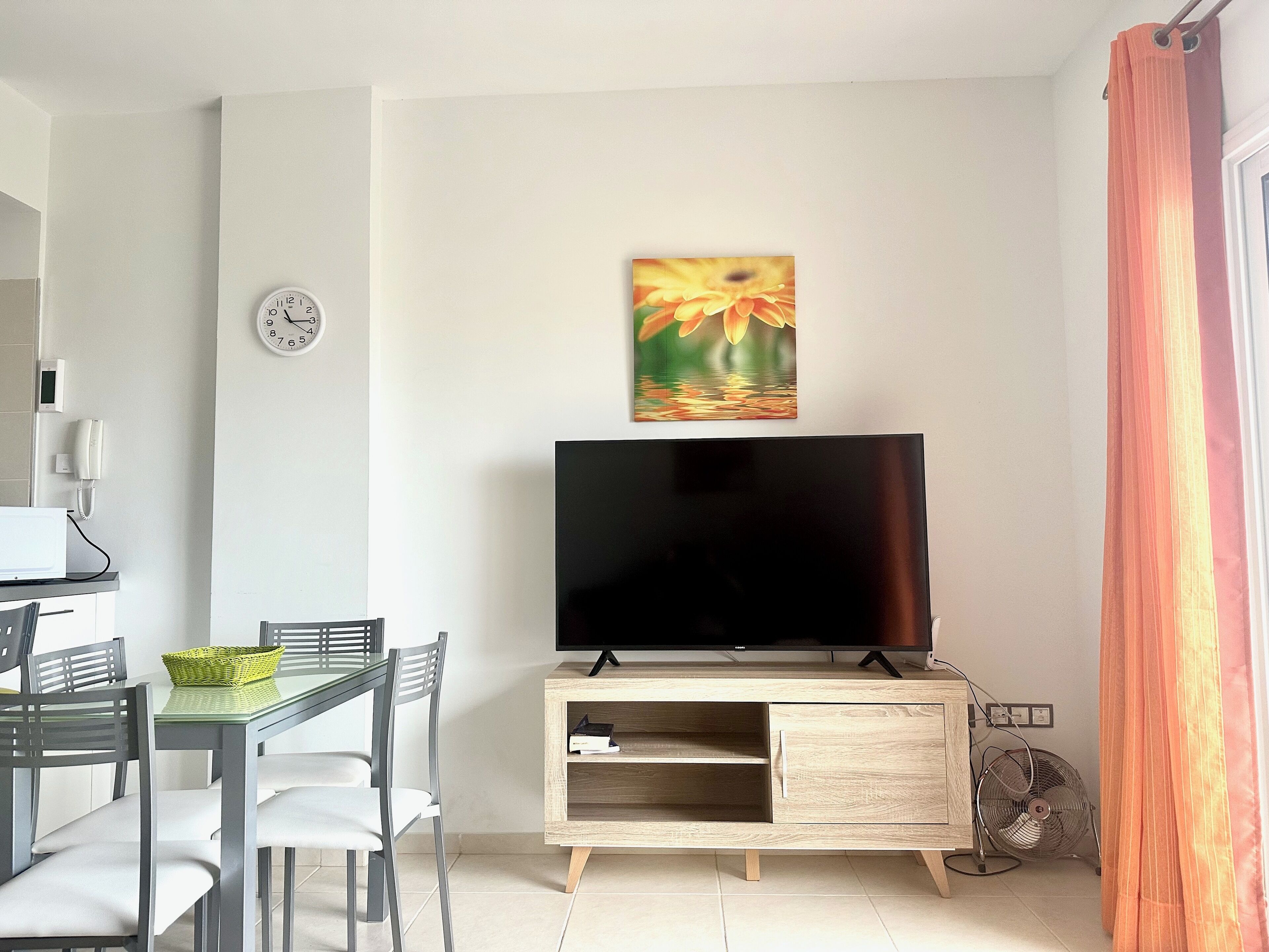 A modern living room with a flat-screen TV on a wooden cabinet, a dining table with chairs, a clock on the wall, and a painting above the TV.
