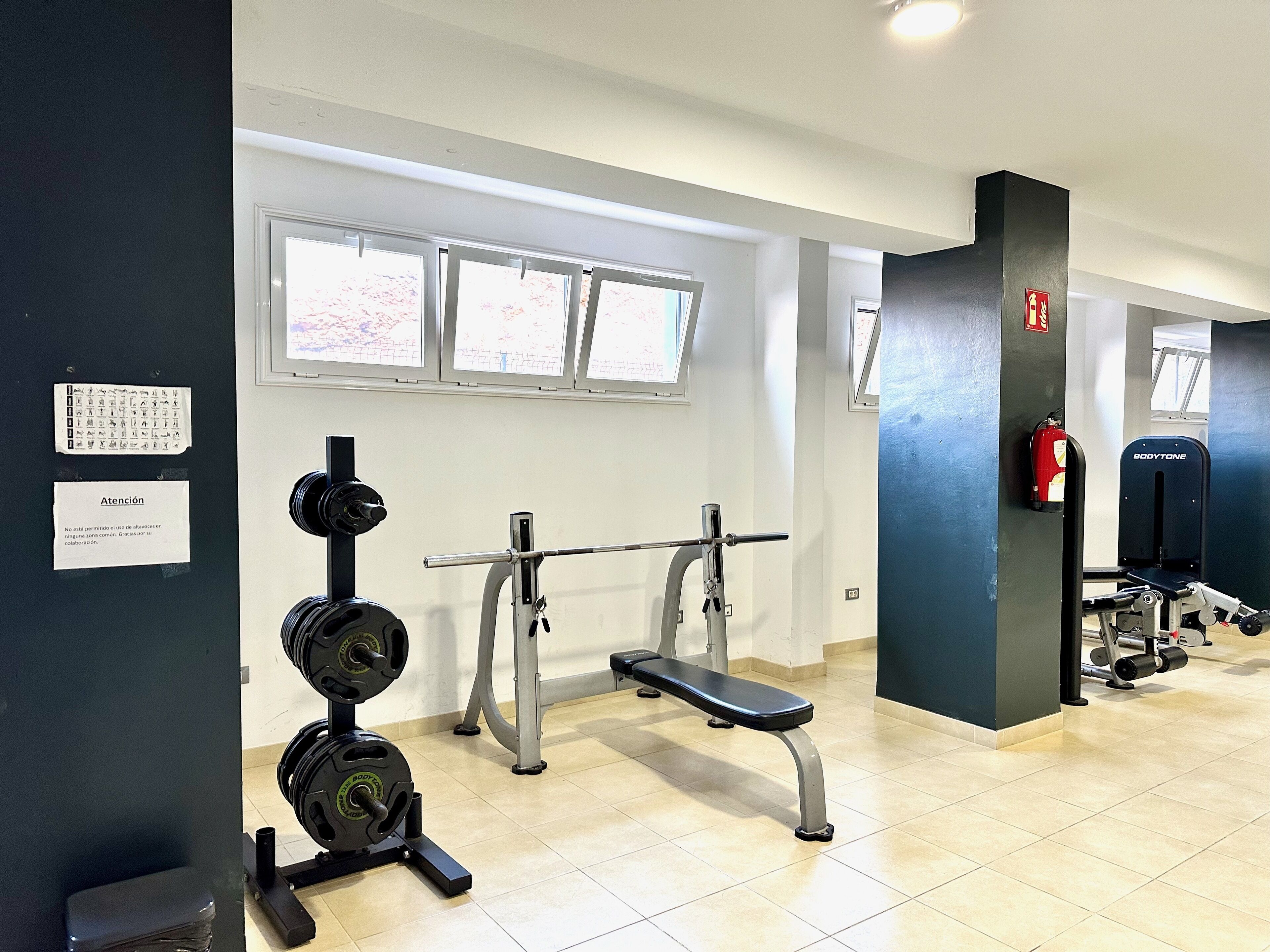 Ein Fitnessraum mit Gewichteinrichtung, einer Hantelbank und einem an der Wand befestigten Feuerlöscher.