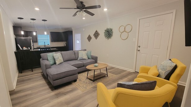 A modern living room with a grey sectional sofa, yellow armchairs, a coffee table, and a ceiling fan. - 2bed/2bath Cottage in Downtown Houma. 30+ day stays welcome. (Houma)