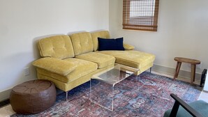 Un canapé d’angle jaune avec un coussin bleu foncé, une table basse en verre transparent, un pouf marron et un tapis à motifs.