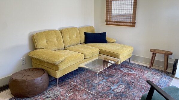 Un sofá seccional amarillo con un cojín azul oscuro, una mesa de centro de vidrio transparente, un reposapiés marrón y una alfombra estampada.