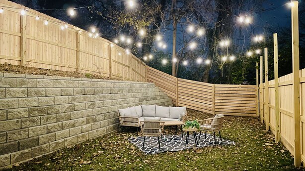 Un jardin avec un mur en pierre, une clôture en bois, des guirlandes lumineuses, un canapé blanc, deux chaises et un tapis à motifs.