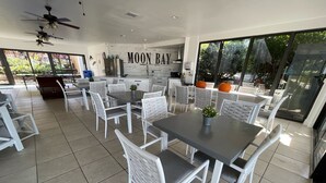 A spacious dining area with white chairs and tables, a kitchen area with stainless steel appliances, and a large window offering a view of the outdoors. - Sunsets and Turtles – Key Largo Moon Bay Condo – Boat Slip/Lift Optional (Key Largo)
