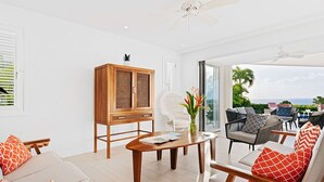 A spacious living area with a wooden cabinet, a white wicker chair, and a dining table. - Incredible villa, beach club access- Whitehaven  (2 bed) (Westmoreland)