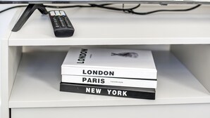 Estante blanco con un control remoto y tres libros titulados 'LONDON', 'PARIS' y 'NEW YORK'.
