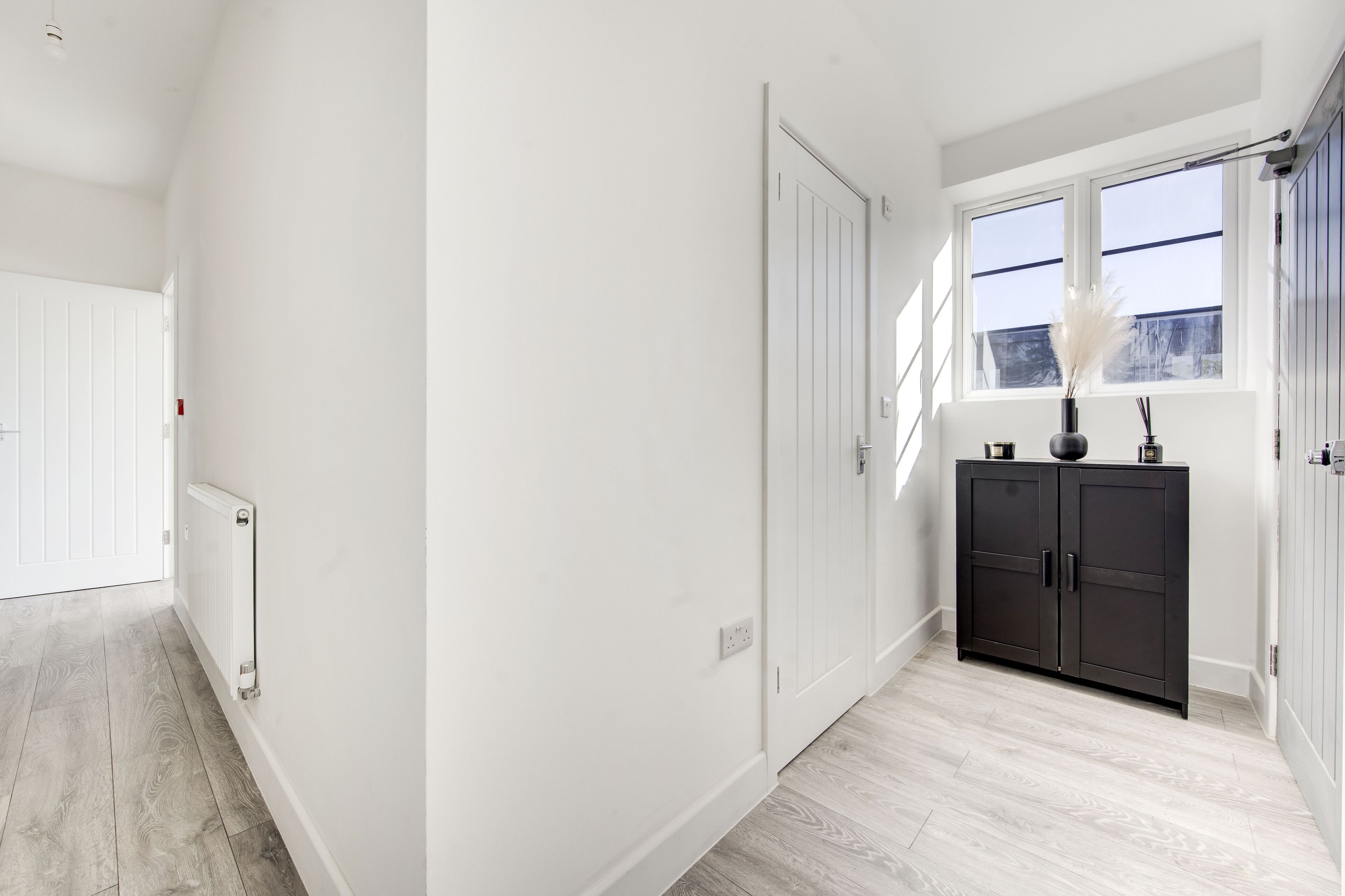 Un couloir aux murs blancs, avec un radiateur et un meuble en bois sombre sur lequel est posé un vase.