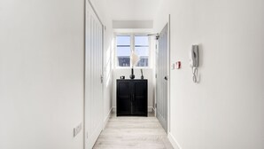 Un pasillo estrecho con paredes blancas y suelo de madera clara. Se observa un mueble negro con un jarrón de plumas de plumeros, una mesita con una vela y un dispositivo fijado a la pared.