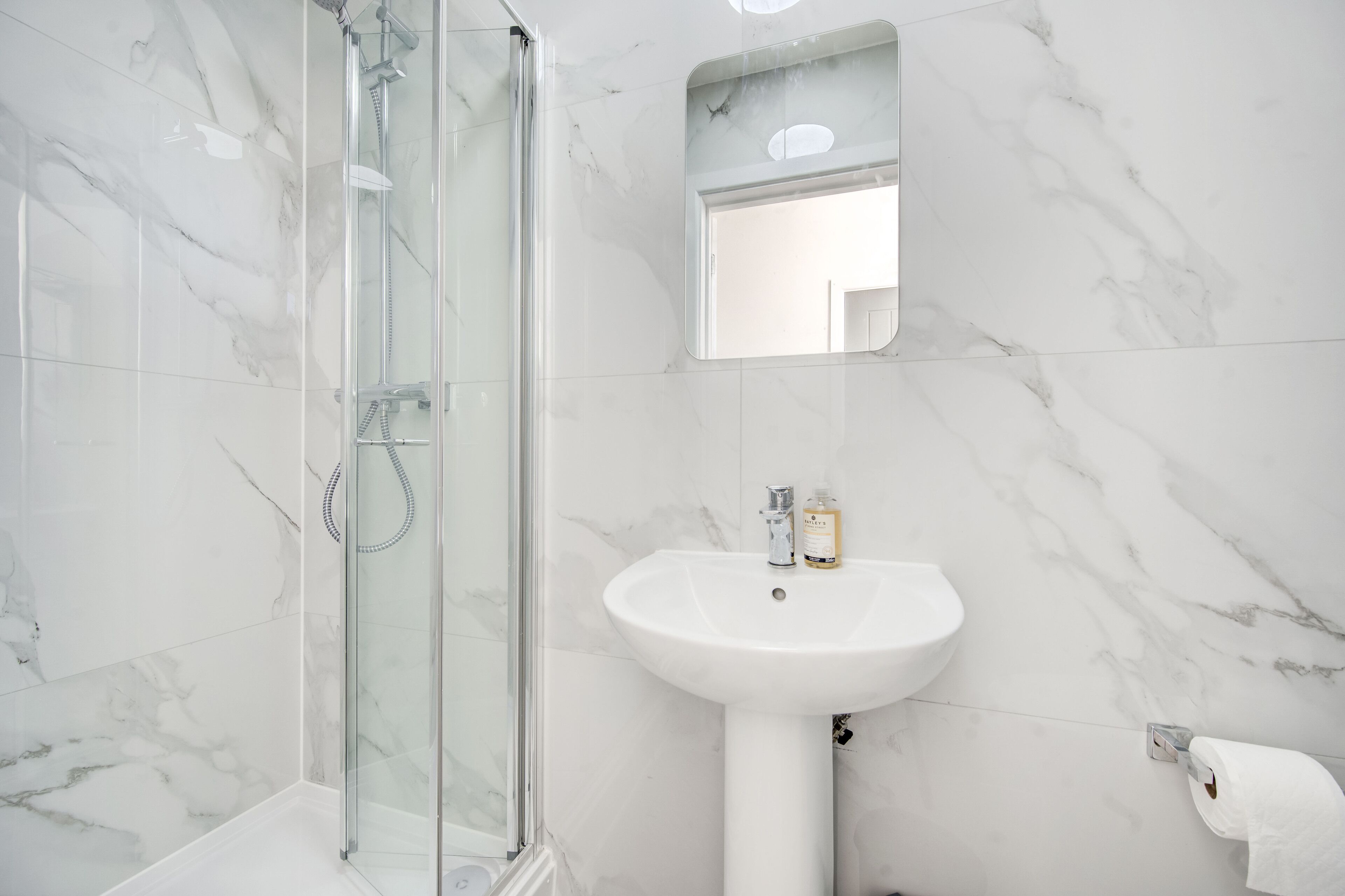 Een badkamer met een douchecabine van glas, een wit wastafeltje en een wand betegeld met marmer.