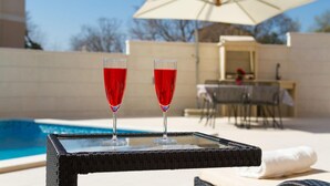 Two glasses of red liquid on a glass table by a pool, with a white umbrella and a dining area in the background. - Awesome home in Liznjan (Liznjan)