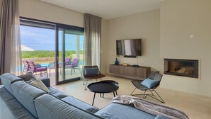 A spacious living room with a large sofa, a flat-screen TV, a fireplace, and a view of a pool and greenery through floor-to-ceiling windows. - Stunning home in Porec with sauna (Porec)