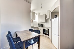A compact kitchen with white cabinets, a black oven, and a white refrigerator. There is a wooden table with blue chairs. - Cosy 2 Bedroom Apartment in Brixton with Wi-Fi and fast route to Central London (London)