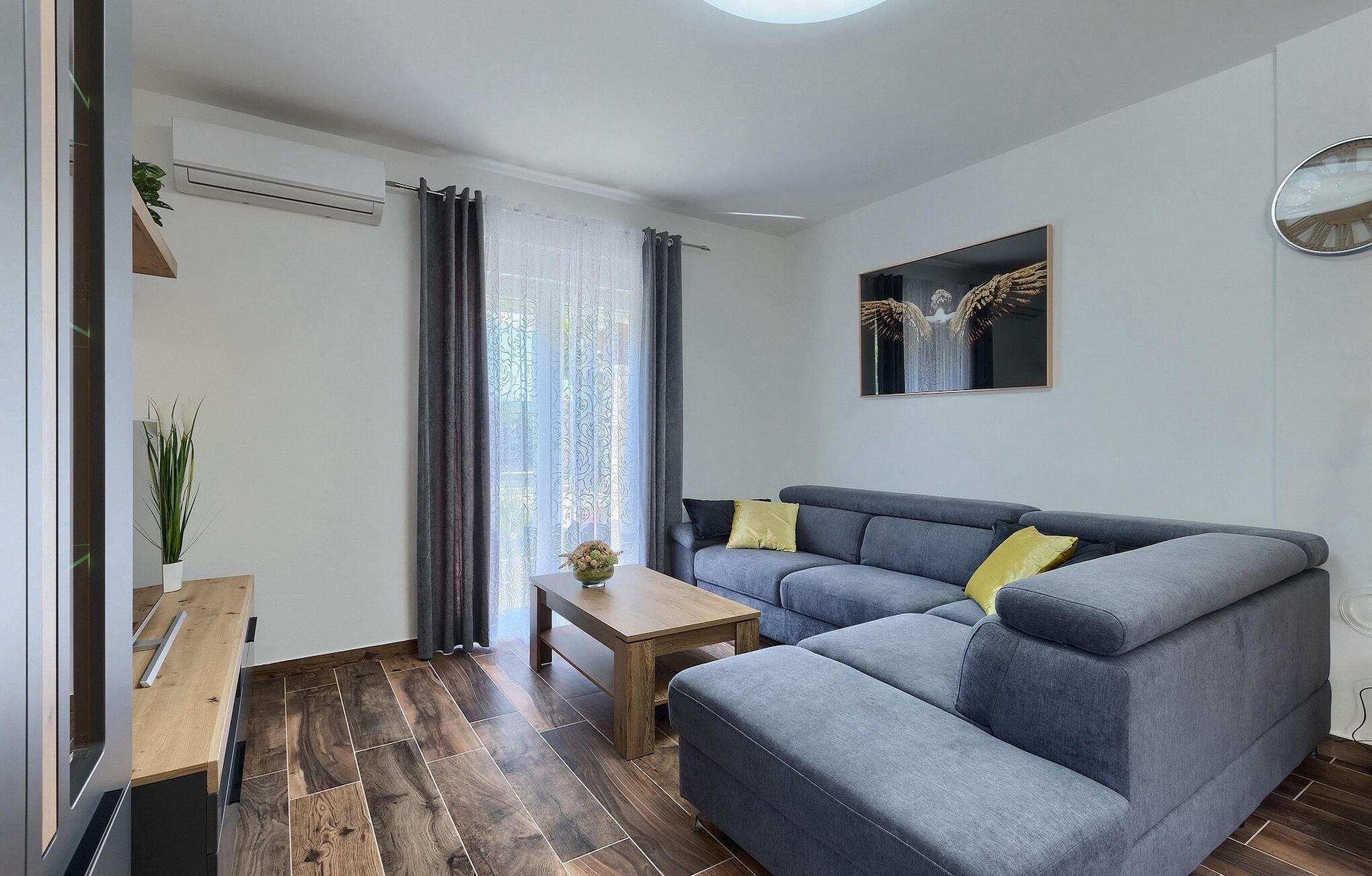 Un salon moderne avec un canapé d’angle gris, une table basse en bois et une petite table d’appoint. On y trouve une fenêtre avec des rideaux, un climatiseur fixé au mur et une œuvre d’art encadrée au-dessus du canapé.