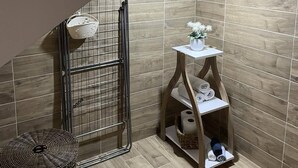 Une salle de bain avec un plancher en bois, un porte-serviettes en métal, un tabouret en osier et une petite étagère en bois où sont posés des serviettes et des articles de toilette.