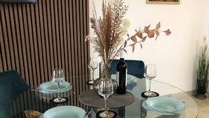 Un coin repas avec une table en verre entourée de chaises recouvertes d’un tissu bleu, un vase contenant des fleurs séchées et une bouteille de vin.