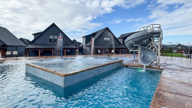 A swimming pool with a water slide and a hot tub, surrounded by buildings with dark roofs and a clear sky.