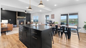 A modern kitchen with a central island, a dining area with a table and chairs, a fireplace, and large windows offering a view.