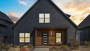 A modern two-story house with a dark exterior, a front porch with two chairs, and a small garden.