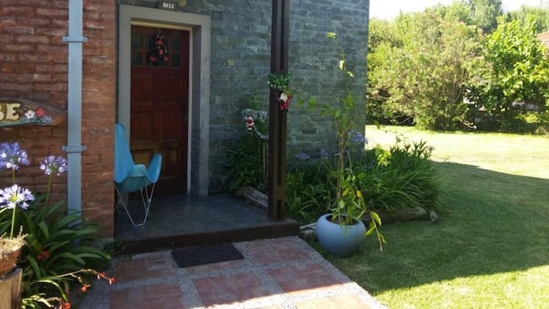 En murstensbygning med en trædør, en lille veranda med stole, og en have med planter og blomster.