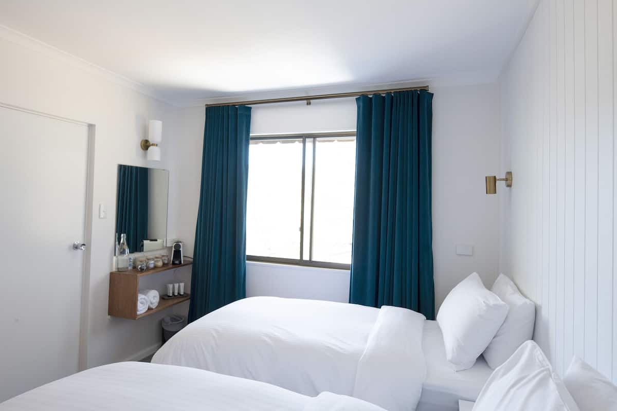 A hotel room with two beds, a window with curtains, a bedside table with a lamp, and a mirror.