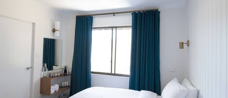 A hotel room with two beds, a window with curtains, a bedside table with a lamp, and a mirror.