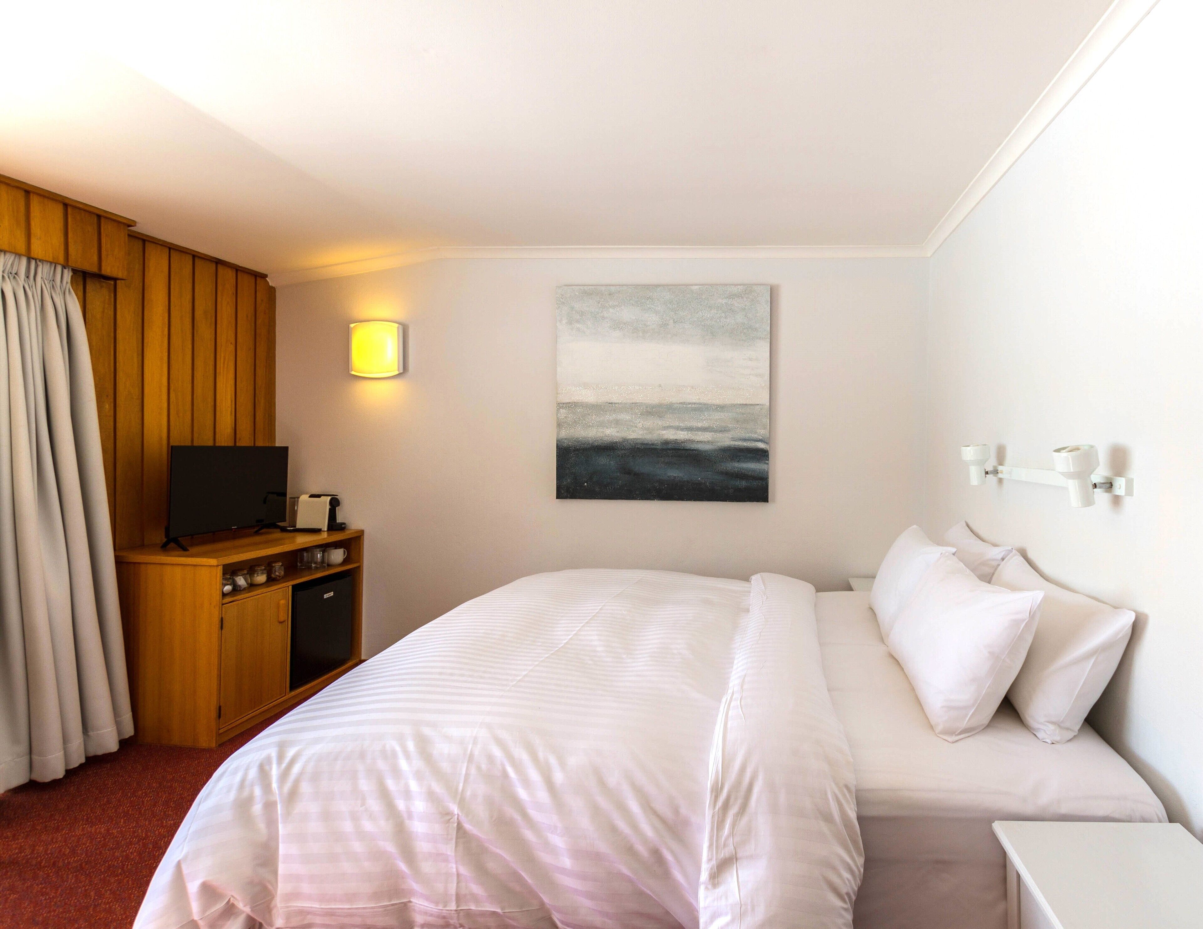 Een hotelkamer met een bed, een televisie op een houten kast, een schilderij aan de muur en een lamp.