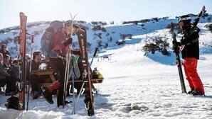 A group of people with skis and ski poles gathered around a wooden bench in a snowy outdoor setting. - Eiger Chalet (Perisher Valley)