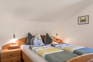Un lit bien fait, recouvert d’une couverture à motifs, une table de chevet en bois avec un tiroir et un luminaire fixé au mur.