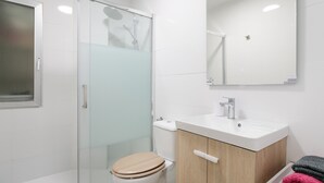 Une salle de bain moderne avec une douche à l’italienne en verre, des toilettes blanches, un meuble-lavabo en bois et un miroir au-dessus du meuble-lavabo.