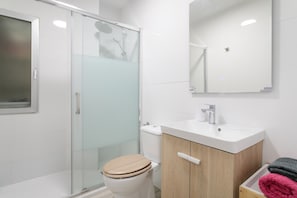 Un baño moderno con cabina de ducha de vidrio, inodoro blanco, tocador de madera con lavamanos y espejo sobre el tocador.