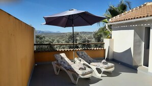 Ein Balkon mit zwei Liegestühlen, einem großen Sonnenschirm und Blick auf eine gebirgige Landschaft.