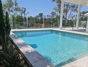 Una piscina rectangular con agua azul cristalina, rodeada por una terraza de baldosas blancas y una valla de cristal.