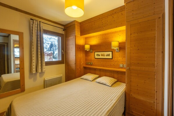 A wooden-paneled bedroom with a bed, a window with curtains, a mirror, and wall-mounted lights.