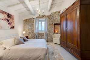 Un dormitorio con pared de piedra, un armario grande de madera, una cama con ropa de cama blanca y una araña.