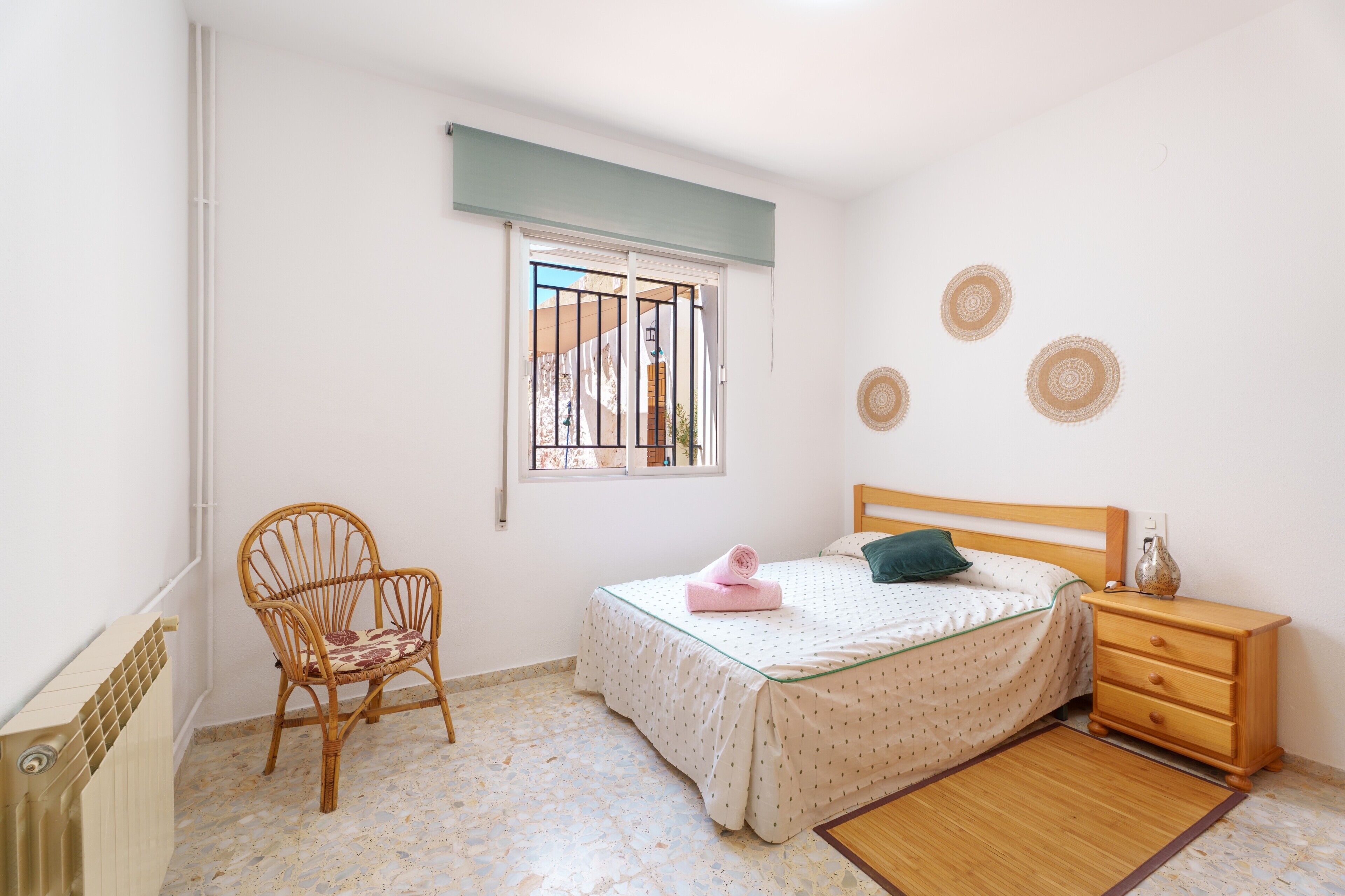 A bedroom with a bed, a wooden nightstand, a chair, a window with bars, and a radiator.