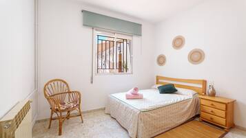 A bedroom with a bed, a wooden nightstand, a chair, a window with bars, and a radiator.