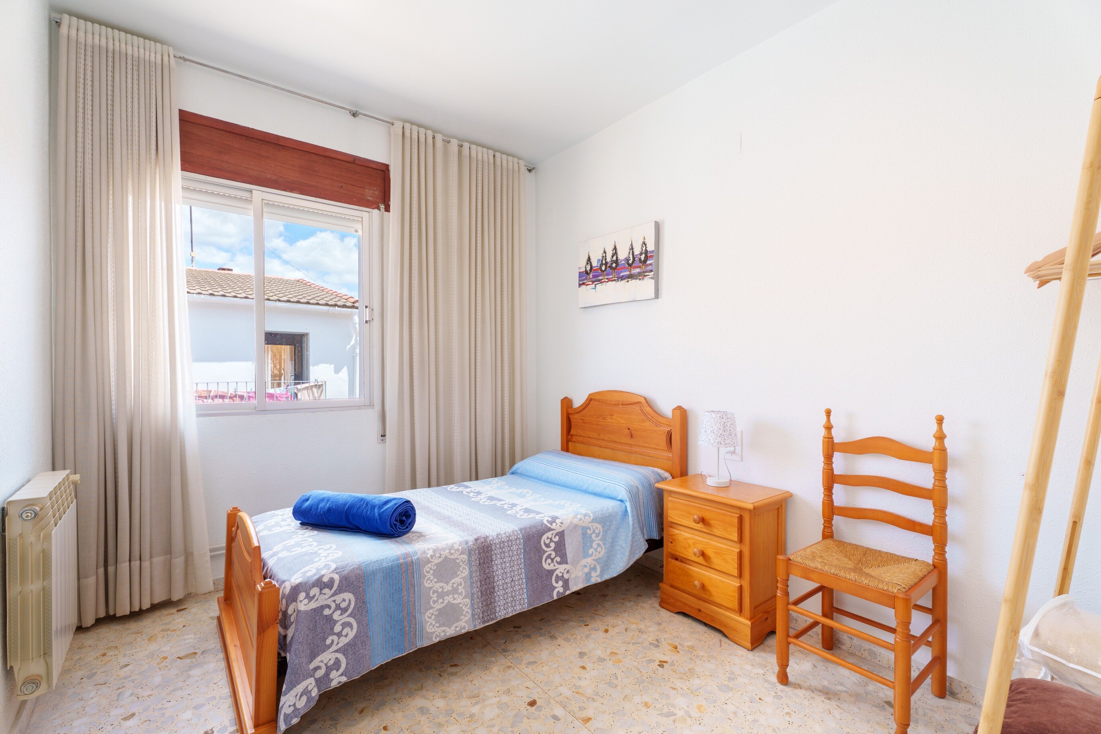 Un dormitorio con cama, una silla de madera, una mesita de noche, un radiador y una ventana con cortinas.
