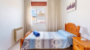Une chambre à coucher comprenant un lit, une table de chevet en bois, un radiateur, une fenêtre avec des rideaux et un tableau accroché au mur.