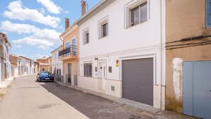 Une rue étroite, bordée de bâtiments des deux côtés, avec une voiture bleue garée à gauche et un portail de garage fermé à droite.