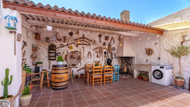 Une terrasse extérieure rustique, équipée d’une machine à laver, de chaises en bois, d’une table et de divers objets suspendus.