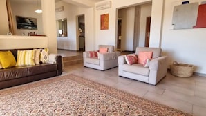 A living room with a brown sofa, two armchairs, a patterned rug, and a hallway leading to a kitchen.
