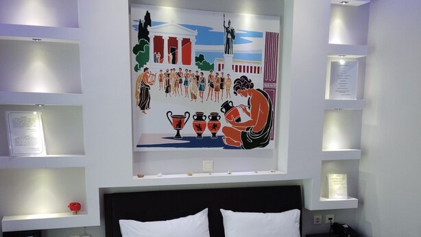 A hotel room with a bed, bedside tables, a nightstand, a wall-mounted artwork, and a shelf with a framed certificate.