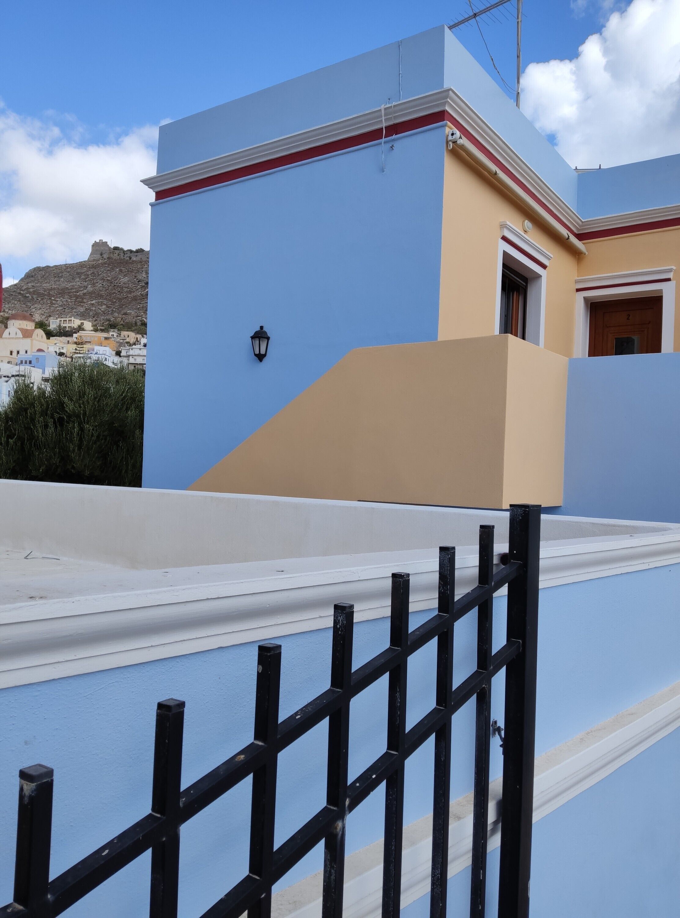 Un edificio con fachada azul y beige, una valla de metal negro y vista a una colina con otros edificios.