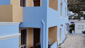 A two-story building with a blue facade, a stone-tiled patio, and a view of a hillside.