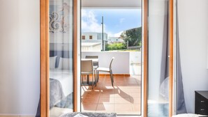 Puerta corredera de vidrio abierta que da a un balcón con piso de baldosas, una mesa, sillas y vistas a edificios y vegetación.